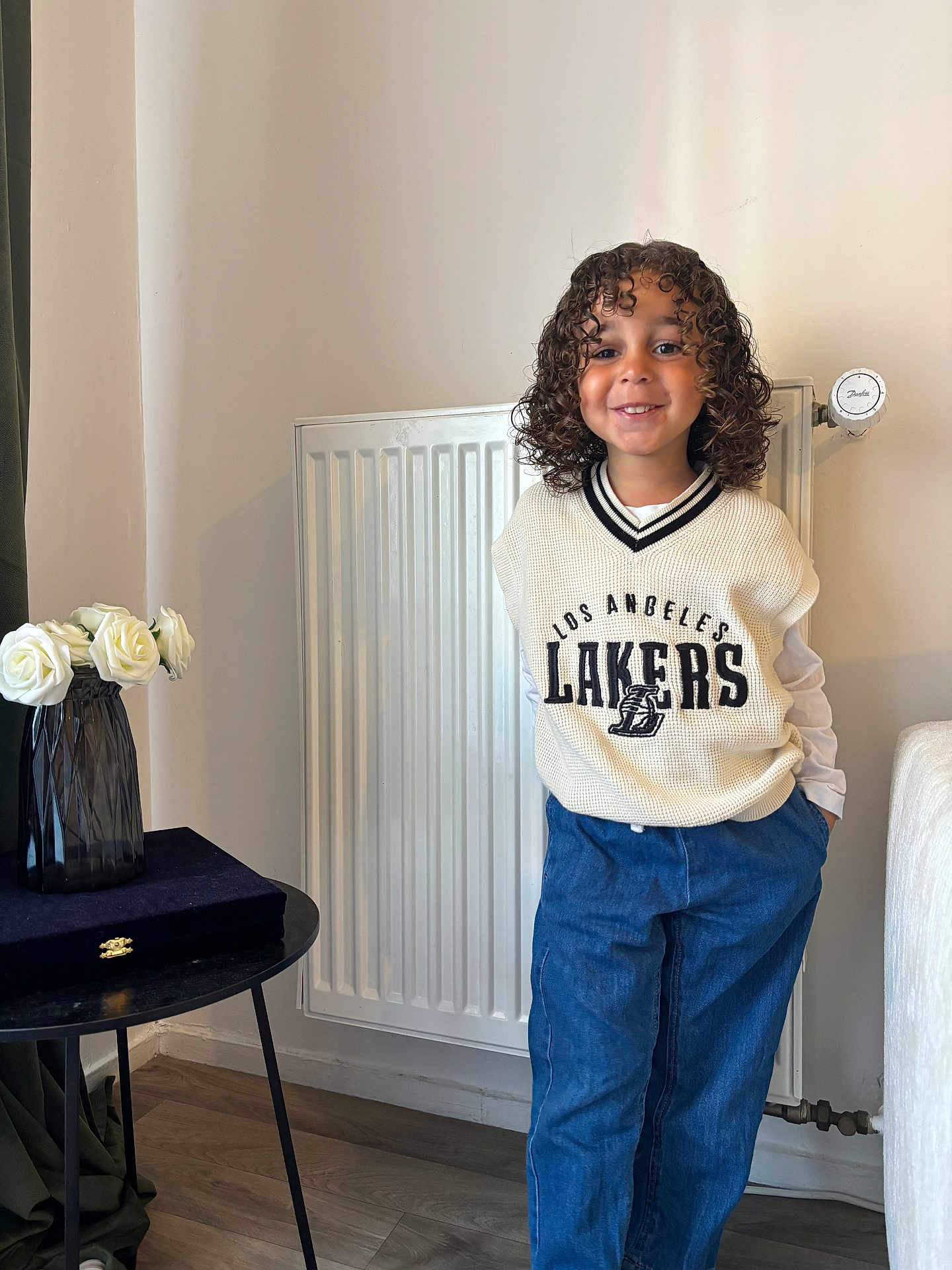 Isaïah a rejoint le concours — aidez-le/la à gagner de superbes lots ! child, curly_hair, smiling, indoor, sweater_vest, blue_pants, hands_in_pockets, radiator, vase, white_roses, table, wooden_floor, casual_clothing, portrait, person, happy, standing, home_interior, decor, fashion