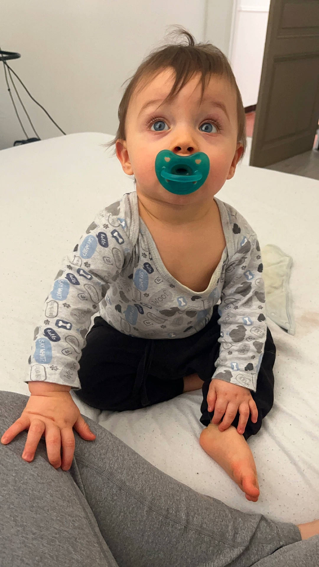 baby, barefoot, bed, black_pants, blanket, blue_eyes, child, curious, cute, door, floor, gray_pants, hand, indoor, infant, long_sleeve_shirt, looking_up, pacifier, person, sitting