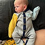 baby, infant, onesie, clothing, person, indoor, couch, window, curious, child, sitting, lap, yellow_sleeve, blue_trim, skin, head, face, button, fabric, cozy