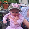 baby, child, hat, pink_onesie, adult_hand, outdoor, garden, plant, flower_pot, table, camouflage, expression, wide_eyes, person, holding, wooden_deck, casual_clothing, nature, summer, portrait