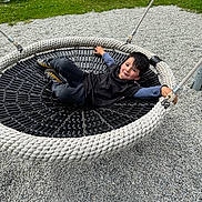 Adam a rejoint le concours — aidez-le/la à gagner de superbes lots ! child, boy, swing, basket_swing, playground, gravel, rope, grass, picnic_table, smiling, black_shirt, sneakers, play, relaxed, outdoor, recreation, portrait, face, seated, metal_post