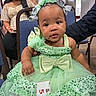 baby, infant, child, portrait, seated, green_dress, sparkly_dress, sequins, bows, hair_bows, festival, number_badge, chair, indoor, adults, background, face, looking_at_camera, formal_event, colorful_clothing