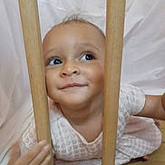 Isabella joined the competition — help win amazing prizes! baby, child, face, wooden_bars, curious, smile, white_dress, patterned_clothing, indoor, floor, hand, eyes, cute, portrait, playful, young_child, skin, head, expression, background