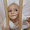 baby, child, face, wooden_bars, curious, smile, white_dress, patterned_clothing, indoor, floor, hand, eyes, cute, portrait, playful, young_child, skin, head, expression, background