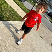 Malekhi is registered to the contest to win money with this photo: toddler, child, sidewalk, curly_hair, red_shirt, black_shorts, white_sneakers, shadow, grass, car, street, residential_area, summer, outdoor, daytime, walking, person, casual_clothing, house, mailbox