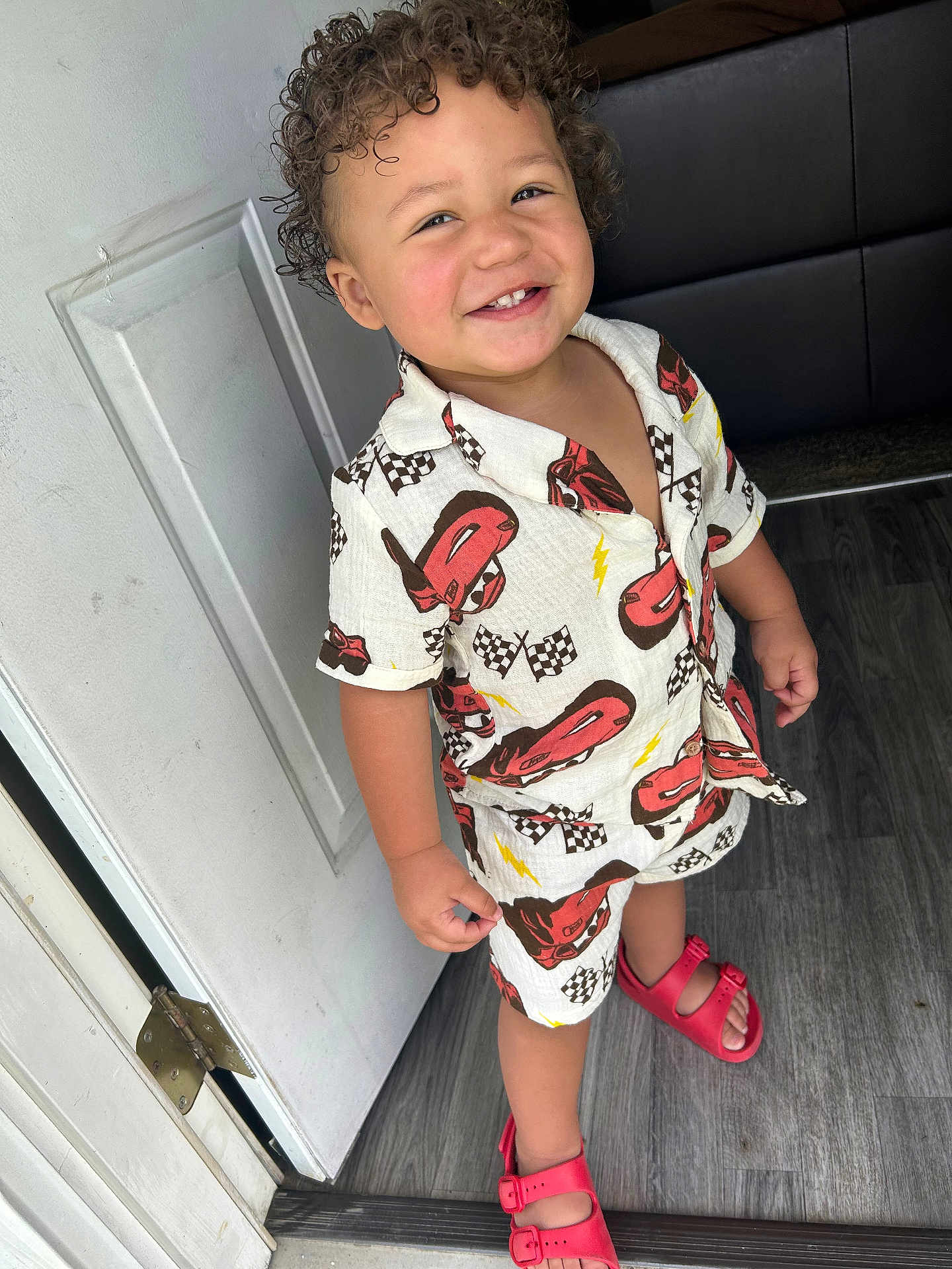 Malekhi joined the competition — help win amazing prizes! toddler, child, curly_hair, smiling, standing, indoors, door, white_door, racing_cars, checkered_flags, shorts, shirt, red_sandals, flooring, happy, cute, clothing, person, portrait, young_child