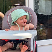 Aleks a rejoint le concours — aidez-le/la à gagner de superbes lots ! baby, child, high_chair, hat, smiling, barefoot, outdoor, window, glass_door, seat, foot, playful, happy, person, infant, sunlight, home, furniture, diaper, curtain