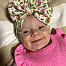 baby, smile, headband, bow, pink_clothing, infant, face, child, cute, apparel, blanket, pattern, portrait, happy, ruffle, fabric, skin, closeup, laying, indoors