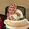 Gracie is registered to the contest to win money with this photo: baby, high_chair, bow, watermelon_pattern, smile, child, indoor, person, clothing, earrings, wood_floor, wall, electrical_outlet, table, seat, playful, cute, infant, tongue_out, happy
