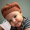 baby, child, infant, portrait, smiling, happy, playful, knit_hat, beanie, striped_shirt, close_up, face, eyes, rosy_cheeks, indoors, bedroom, pillow, home, soft_lighting, photograph