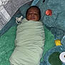 newborn, baby, swaddled, blanket, sleeping, plush_toy, stuffed_animal, crib, mesh_sides, pacifier, soft_texture, green, blue, orange, infant, cute, resting, child, indoors, cozy