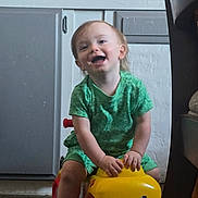 Kayn is registered to the contest to win money with this photo: toddler, child, toy_car, indoor, green_clothing, happy, smiling, playing, floor, cabinet, wooden_floor, yellow_toy, red_wheels, person, young_child, cute, fun, sitting, home, furniture