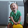 toddler, child, toy_car, indoor, green_clothing, happy, smiling, playing, floor, cabinet, wooden_floor, yellow_toy, red_wheels, person, young_child, cute, fun, sitting, home, furniture