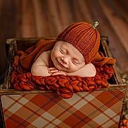 James is registered to the contest to win money with this photo: baby, sleeping, orange_hat, knitted_hat, wooden_box, blanket, cozy, infant, newborn, smile, resting, cute, plaid_pattern, fall_theme, wood_floor, child, portrait, indoors, peaceful, warm_colors