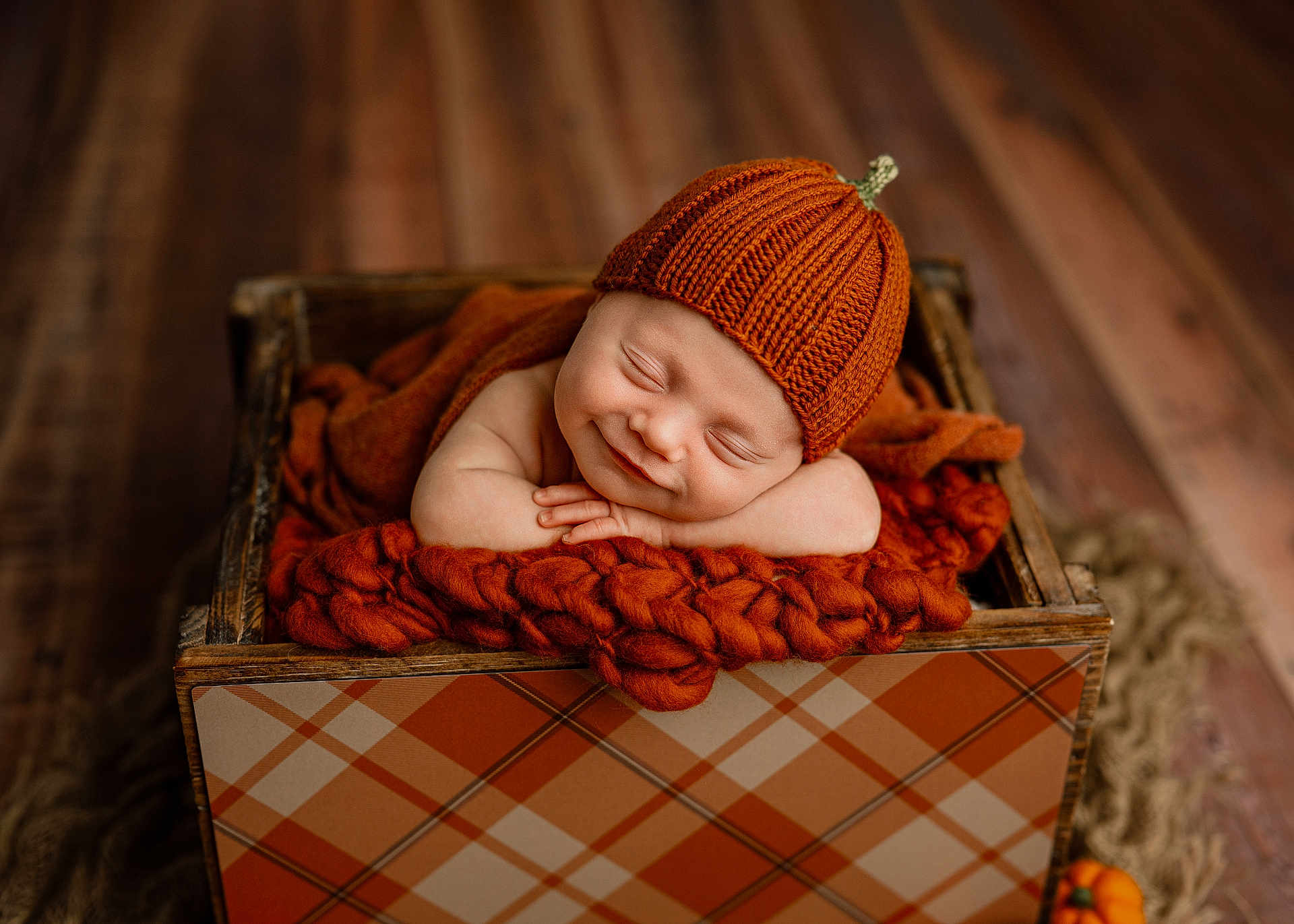 James is registered to the contest to win money with this photo: baby, sleeping, orange_hat, knitted_hat, wooden_box, blanket, cozy, infant, newborn, smile, resting, cute, plaid_pattern, fall_theme, wood_floor, child, portrait, indoors, peaceful, warm_colors