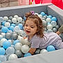 toddler, child, ball_pit, playroom, colorful, curly_hair, scrunchie, purple_clothing, blue_balls, white_balls, silver_balls, padded_edges, indoor, happy, play, fun, smiling, toy, soft_furniture, cushion