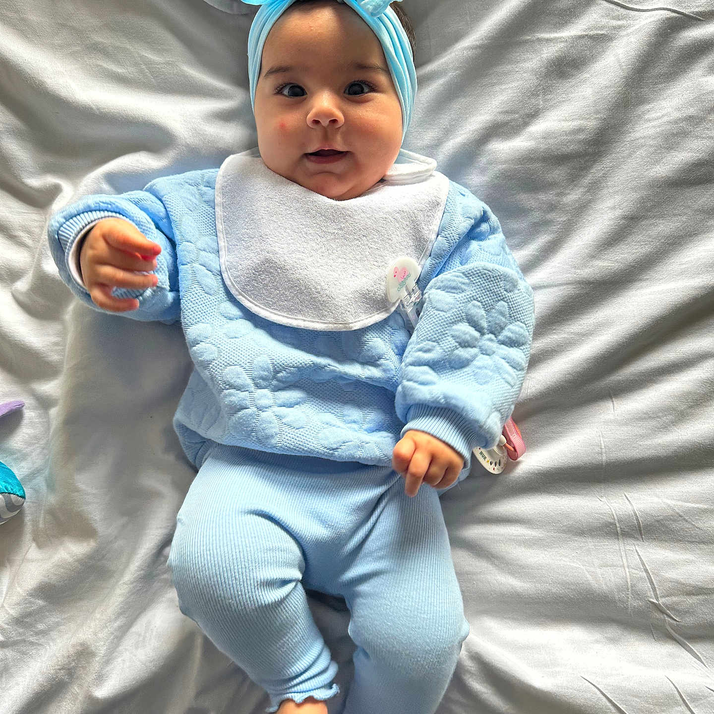 Amyra is registered to the contest to win money with this photo: baby, bed, blue_clothing, child, comfortable, cozy, cute, hand, headband, indoor, infant, knitwear, lying_down, person, playful, portrait, small_feet, smiling, soft_light, white_sheet