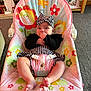 baby, infant, child, fisher_price, baby_chair, headband, black_and_white_clothing, hand_in_mouth, colorful, flower_pattern, bee_pattern, indoor, carpet, furniture, photo_frame, living_room, cute, seated, person, smiling