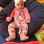 Bonnie-Rae is registered to the contest to win money with this photo: baby, child, pink_clothing, hat, bow, bean_bag, socks, indoor, floor, carpet, adult_arm, wooden_floor, patterned_pants, cute, seated, person, cozy, home, casual, relaxed