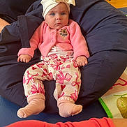 Bonnie-Rae is registered to the contest to win money with this photo: baby, child, pink_clothing, hat, bow, bean_bag, socks, indoor, floor, carpet, adult_arm, wooden_floor, patterned_pants, cute, seated, person, cozy, home, casual, relaxed