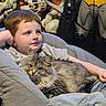 Lynx is registered to the contest to win money with this photo: boy, cat, armchair, dinosaur_toys, batman_figure, gray_cat, child, relaxed, indoor, furniture, soft_fabric, toy, plush, casual_clothing, portrait, pet, mammal, cute, home, comfort