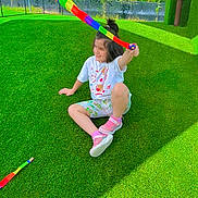 Aliza is registered to the contest to win money with this photo: child, outdoor, sitting, smiling, foam_toy, colorful, green_grass, artificial_turf, playground, backyard, fence, trees, sunlight, shadows, summer, casual_clothing, pink_shoes, t_shirt, shorts, portrait