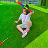 child, outdoor, sitting, smiling, foam_toy, colorful, green_grass, artificial_turf, playground, backyard, fence, trees, sunlight, shadows, summer, casual_clothing, pink_shoes, t_shirt, shorts, portrait