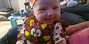 Noelle is registered to the contest to win money with this photo: baby, infant, smiling, headband, turkey_patch, turkey_costume, autumn_print, living_room, couch, adult_hand, holding, hair_spikes, door, toy_table, pillow, blanket, festive, pumpkin, person, indoor