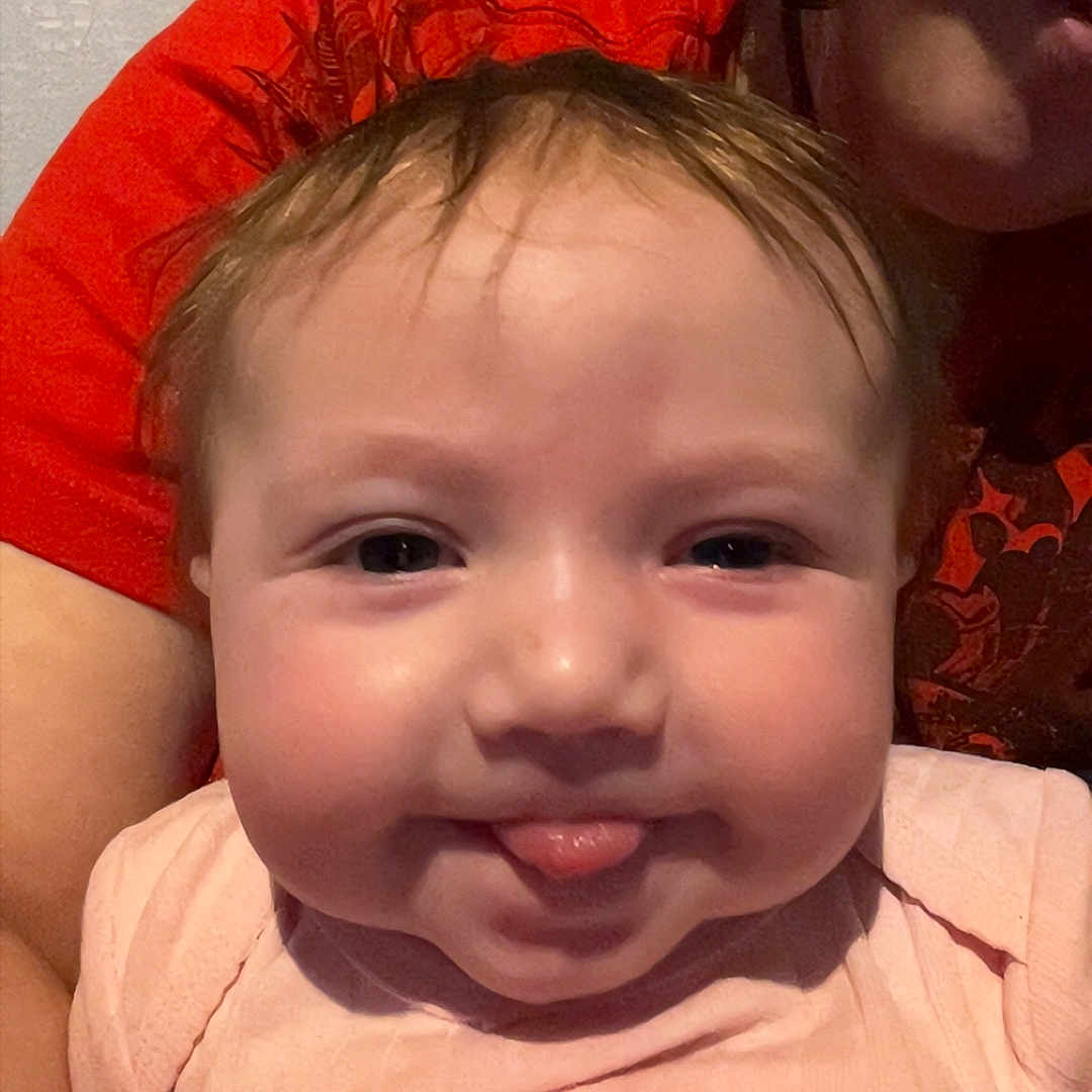 Noelle joined the competition — help win amazing prizes! adult_partial, baby, child, chubby_cheeks, closeup, cute, eyes, family, hair, holding, indoor, infant, person, pink_clothing, portrait, red_shirt, skin, smile, tongue_out, wall