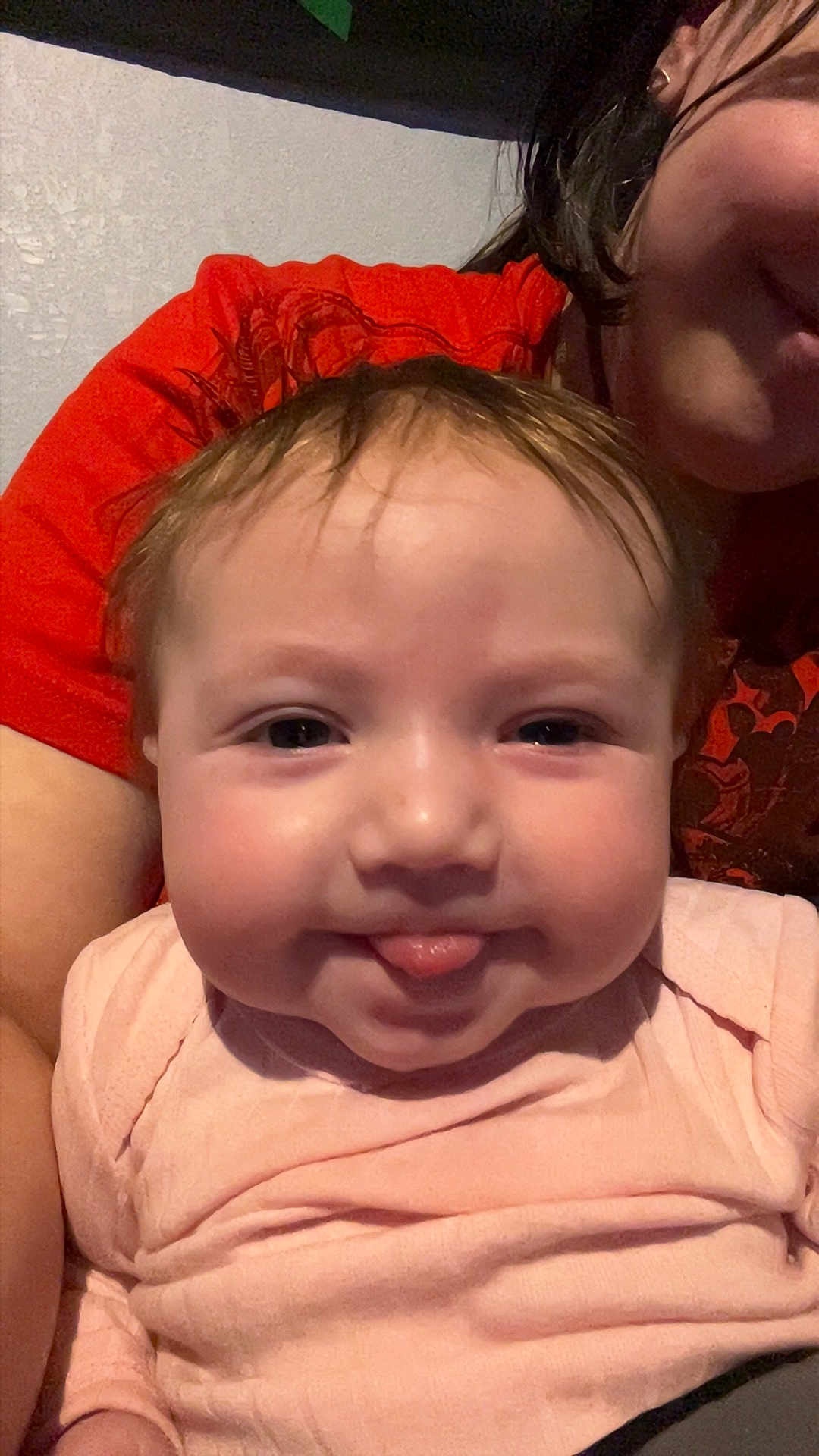 Noelle joined the competition — help win amazing prizes! baby, infant, child, tongue_out, chubby_cheeks, smile, portrait, closeup, person, adult_partial, red_shirt, pink_clothing, indoor, hair, eyes, skin, holding, family, cute, wall