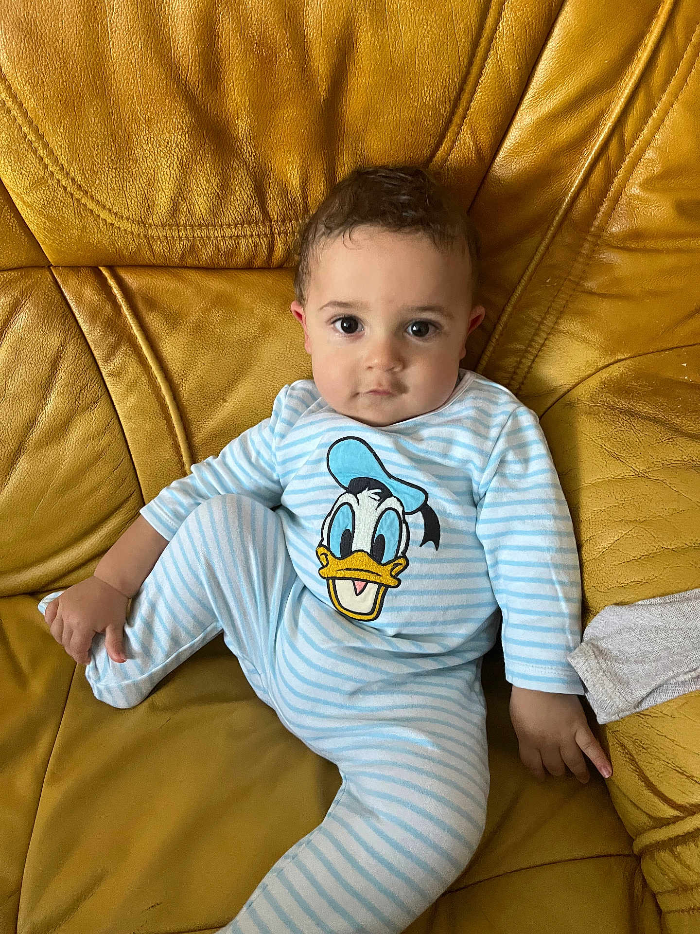 Aiden participe au concours pour gagner de l'argent avec cette photo : baby, child, onesie, donald_duck, striped_clothing, couch, leather, yellow, indoor, portrait, sitting, cute, young_child, face, expression, hand, foot, relaxed, comfortable, infant