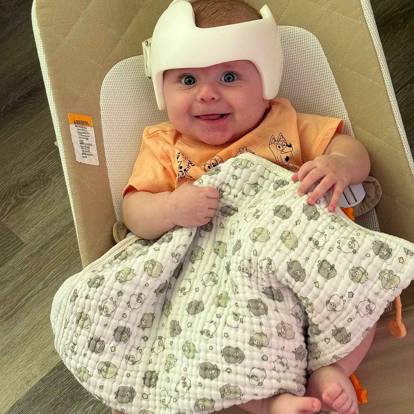 Daniel is registered to the contest to win money with this photo: animal_print, baby, blanket, chair, child, comfort, cushioned_seat, cute, foot, hand, happy, helmet, indoor, infant, orange_shirt, person, protective_gear, resting, smiling, wooden_floor