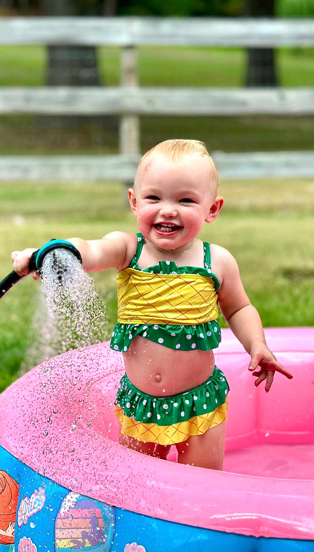 Mary-Alice joined the competition — help win amazing prizes! toddler, child, smiling, water, hose, swimsuit, pool, inflatable_pool, outdoor, summer, playtime, grass, fence, happy, splashing, cute, sunny, water_spray, fun, baby