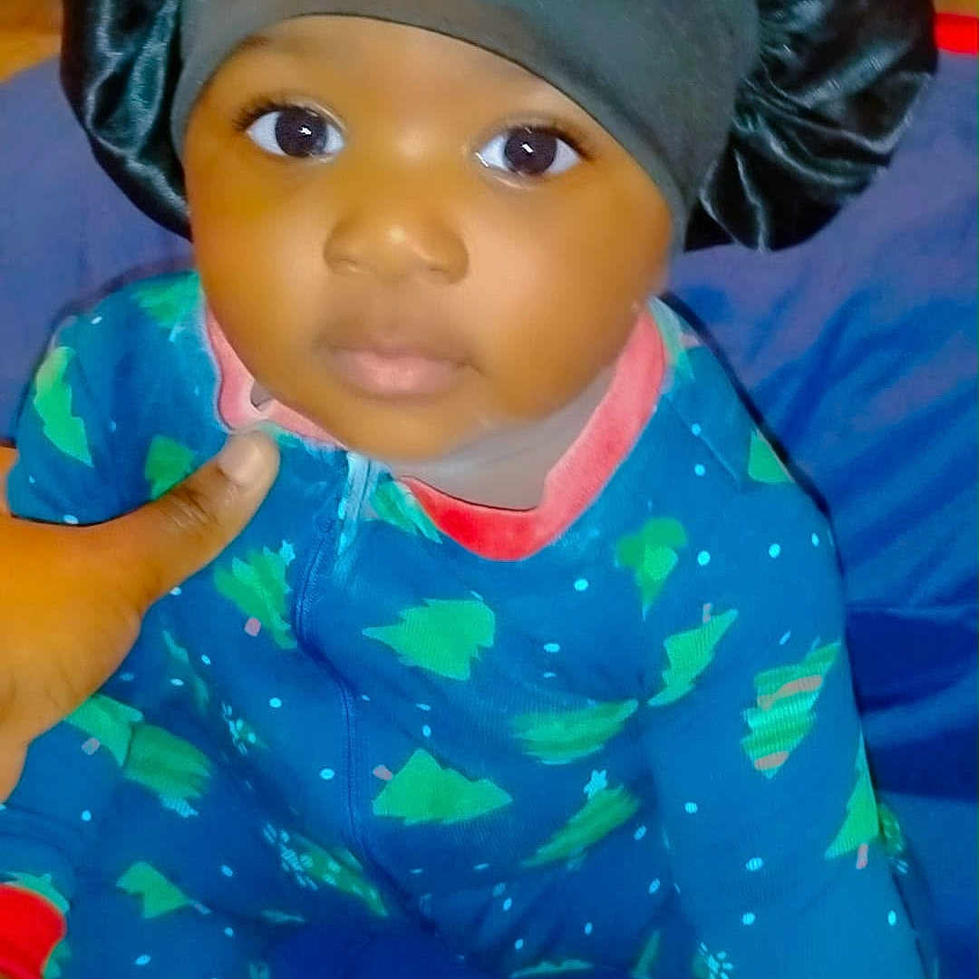 Xamira is registered to the contest to win money with this photo: baby, child, infant, onesie, blue_clothing, christmas_tree_pattern, black_bonnet, satin_bonnet, sitting, hand, indoor, portrait, cute, looking_at_camera, young_child, soft_lighting, close_up, person, footwear, white_sock