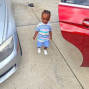 Quadir is registered to the contest to win money with this photo: child, smiling, outdoor, driveway, car, silver_car, red_car, polo_shirt, shorts, white_shoes, braided_hair, concrete, garage_door, bracelet, necklace, person, standing, casual_clothing, daylight, happy