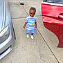 child, smiling, outdoor, driveway, car, silver_car, red_car, polo_shirt, shorts, white_shoes, braided_hair, concrete, garage_door, bracelet, necklace, person, standing, casual_clothing, daylight, happy