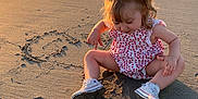 Julia participe au concours pour gagner de l'argent avec cette photo : toddler, child, beach, sand, sunset, shadow, ocean, water, waves, dress, white_shoes, footwear, sitting, heart_drawing, sunlight, outdoor, nature, curly_hair, play, person