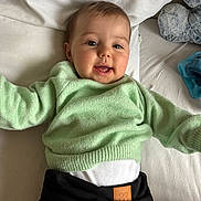 Melya participe au concours pour gagner de l'argent avec cette photo : baby, child, smiling, green_sweater, black_pants, bed, pillow, plush_toy, happy, face, infant, cute, indoor, person, young_child, soft_toys, clothing, lying_down, portrait, cozy