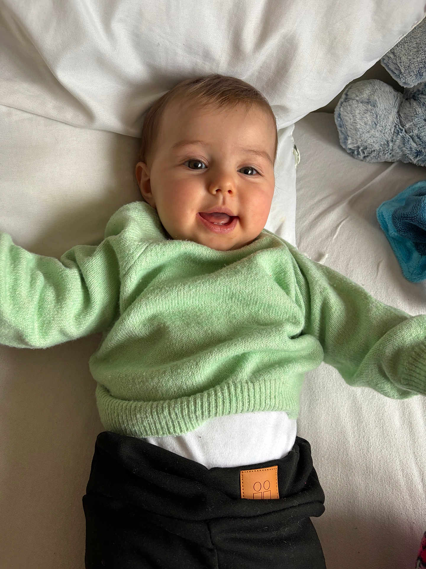 Melya participe au concours pour gagner de l'argent avec cette photo : baby, child, smiling, green_sweater, black_pants, bed, pillow, plush_toy, happy, face, infant, cute, indoor, person, young_child, soft_toys, clothing, lying_down, portrait, cozy