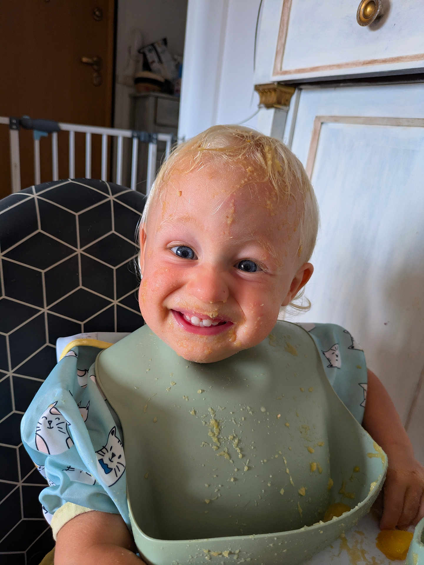 Johan a rejoint le concours — aidez-le/la à gagner de superbes lots ! baby_gate, bib, blond_hair, blue_eyes, child, clothing, cute, feeding, food, happy, high_chair, indoor, infant, kitchen, meal_time, messy_face, person, smile, table, toddler