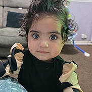 Mina joined the competition — help win amazing prizes! toddler, child, curly_hair, big_eyes, black_clothing, beige_clothing, indoor, living_room, sofa, carpet, wallpaper, portrait, person, cute, young_child, curious, baby, fashion, home, closeup