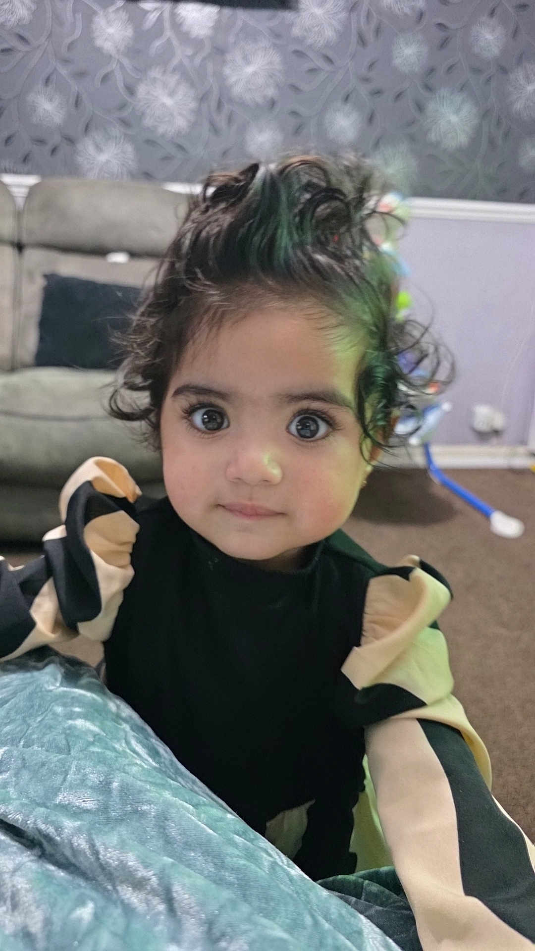 Mina joined the competition — help win amazing prizes! toddler, child, curly_hair, big_eyes, black_clothing, beige_clothing, indoor, living_room, sofa, carpet, wallpaper, portrait, person, cute, young_child, curious, baby, fashion, home, closeup