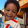 child, toddler, smiling, curly_hair, colorful_clothing, outdoor, cafe, table, chair, building, awning, sky, happy, face, person, daylight, portrait, cute, urban, casual
