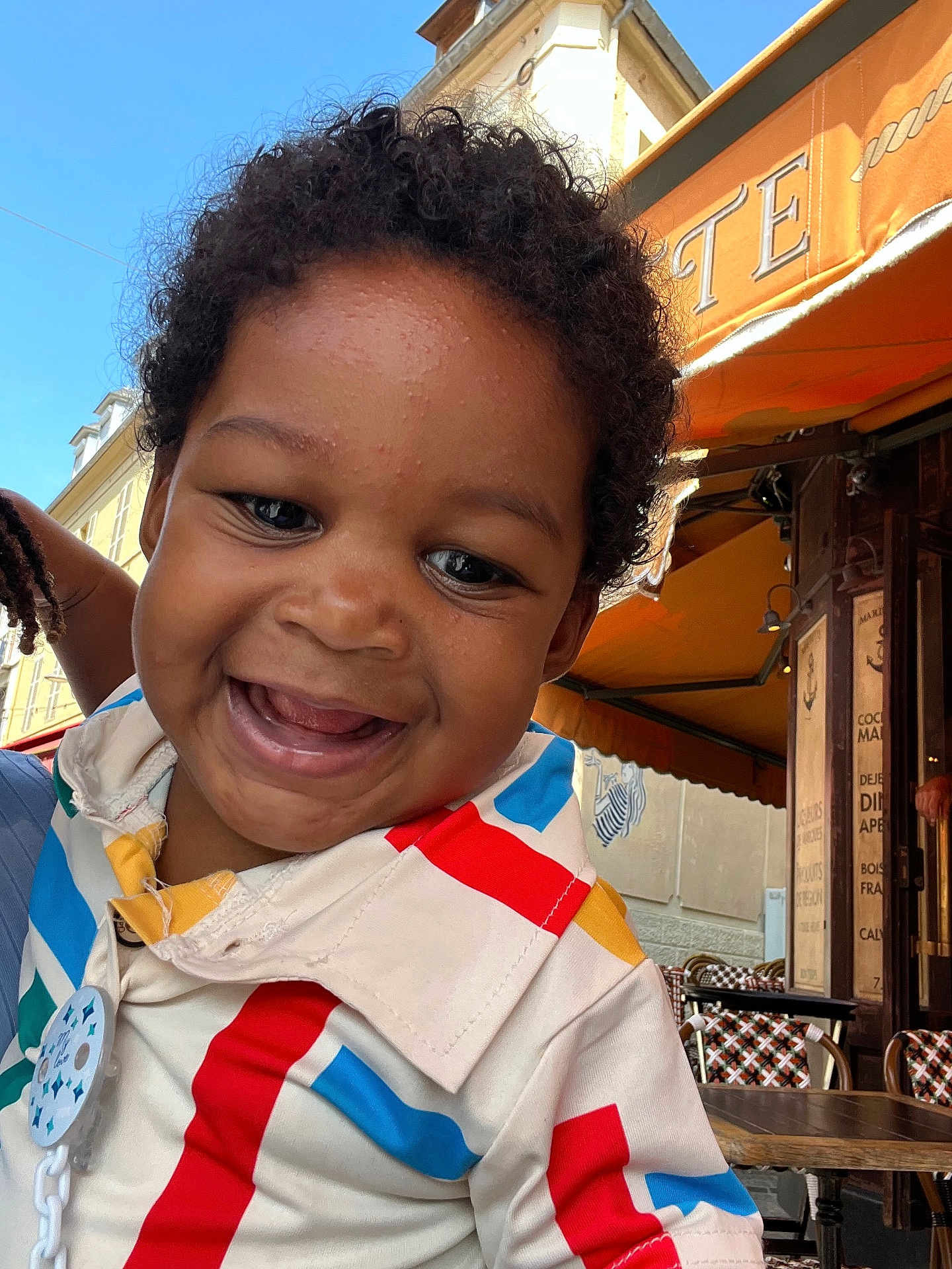 Azhary a rejoint le concours — aidez-le/la à gagner de superbes lots ! child, toddler, smiling, curly_hair, colorful_clothing, outdoor, cafe, table, chair, building, awning, sky, happy, face, person, daylight, portrait, cute, urban, casual