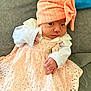 baby, infant, peach_dress, lace, hat, bow, cute, small_hands, couch, gray_background, white_sleeves, portrait, child, sitting, indoors, closeup, soft_lighting, expression, person, newborn