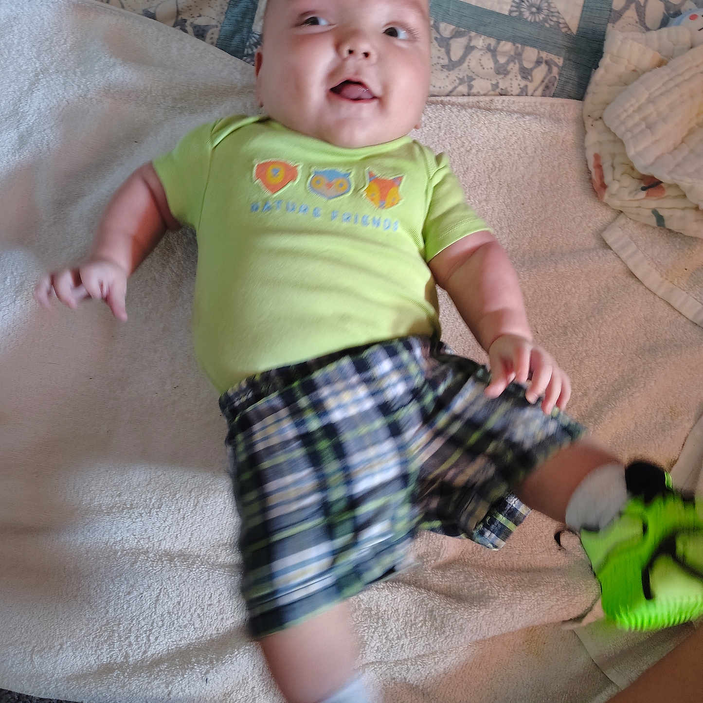 Aleister joined the competition — help win amazing prizes! baby, carpet, child, clothing, cute, footwear, green_shoes, happy, home, indoor, infant, laying_down, motion_blur, patterned_quilt, person, plaid_shorts, shirt, smiling, towel, young_child