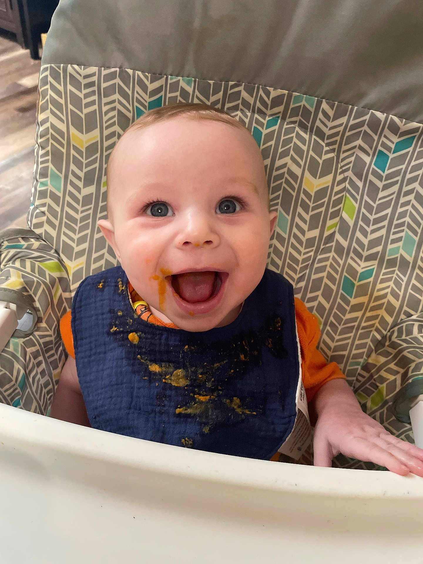 Dominick is registered to the contest to win money with this photo: baby, child, high_chair, bib, messy, smiling, happy, face, blue_eyes, feeding, food_stains, indoor, patterned_fabric, cute, infant, hand, clothing, expression, person, cheerful