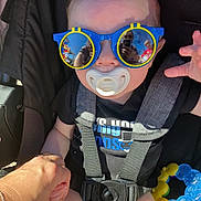 Dominick joined the competition — help win amazing prizes! baby, sunglasses, pacifier, child, reflection, hand, black_shirt, car_seat, harness, toy, blue, yellow, outdoor, person, infant, cute, holding_hand, smile, casual, sunlight