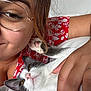 person, cat, glasses, hand, paw, face, bed, red_clothing, floral_pattern, skin, nail_polish, pet, relaxation, close_up, indoor, white_bed, cute, affection, cozy, smile