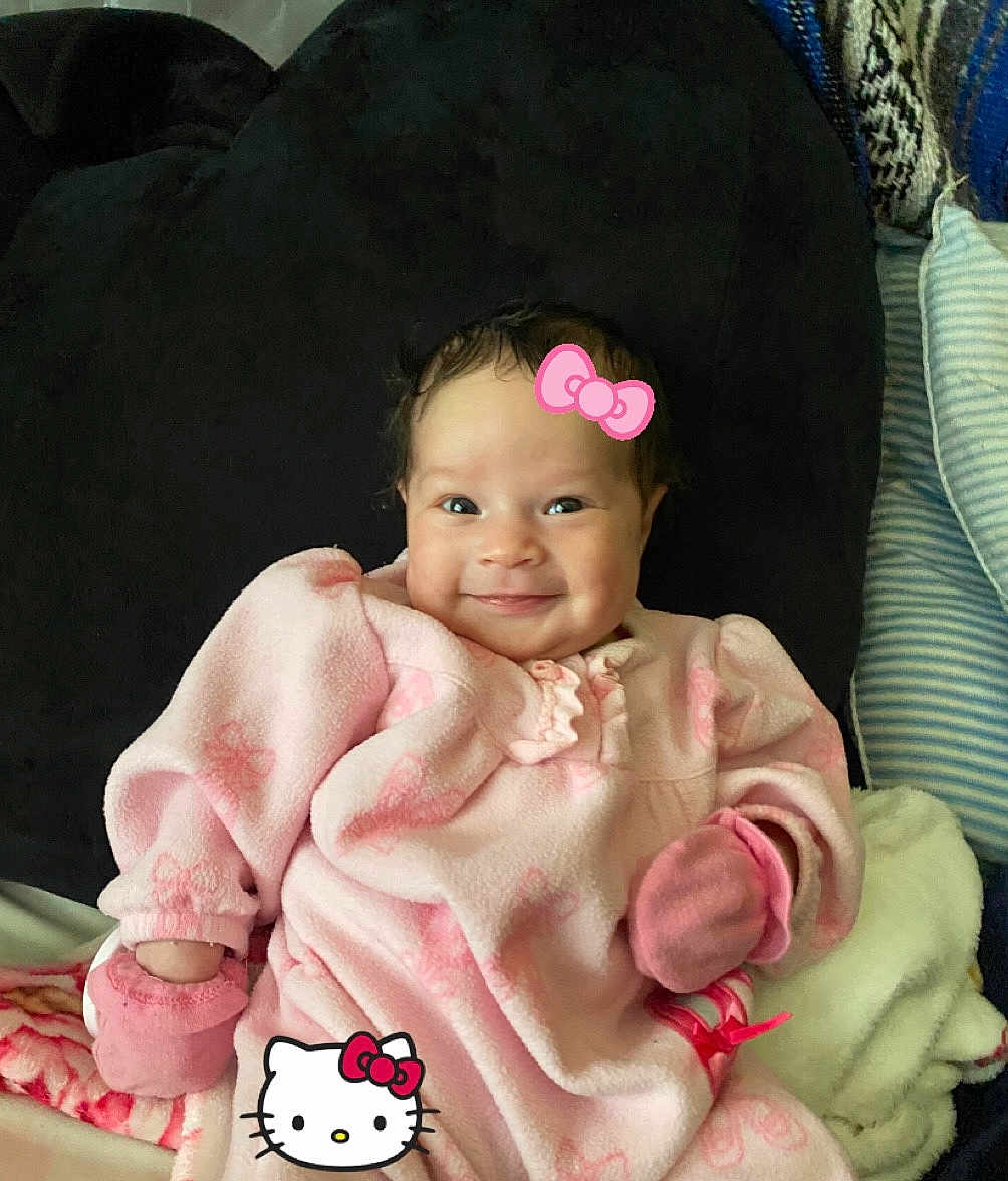 Amora is registered to the contest to win money with this photo: baby, infant, pink_clothing, mittens, smile, cute, cozy, blanket, cushion, indoors, lying_down, face, head, child, soft_texture, hello_kitty, graphic_overlay, happy, portrait, warm
